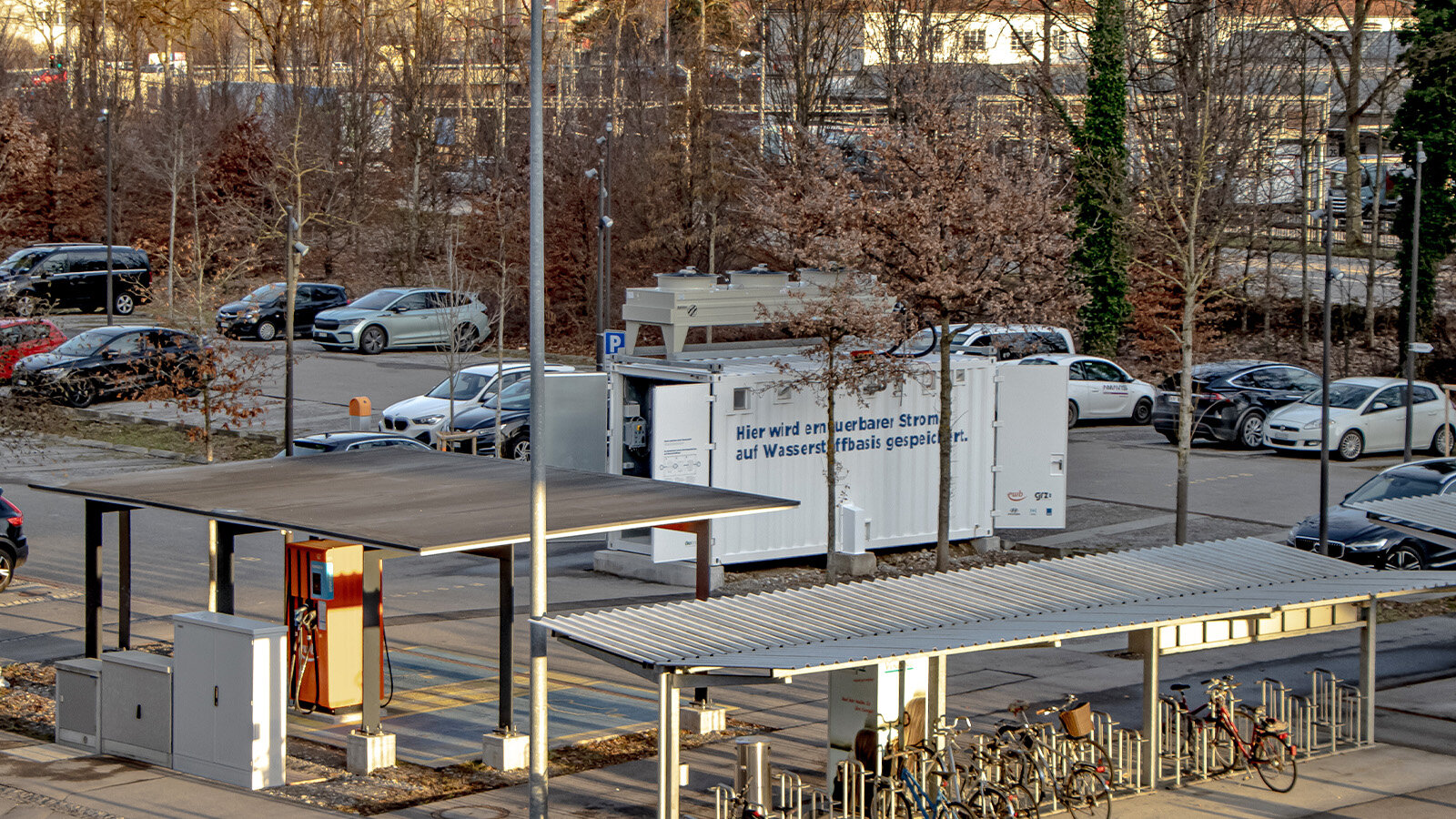 Zu sehen ist aus der Ferne ein Wasserstoffcontainer mit einer blauen Aufschrift, die jedoch nicht lesbar ist. Der Container steht mitten auf einem Parkplatz, umgeben von einigen Autos und Bäumen. Im Vordergrund befindet sich ein Fahrradstellplatz.