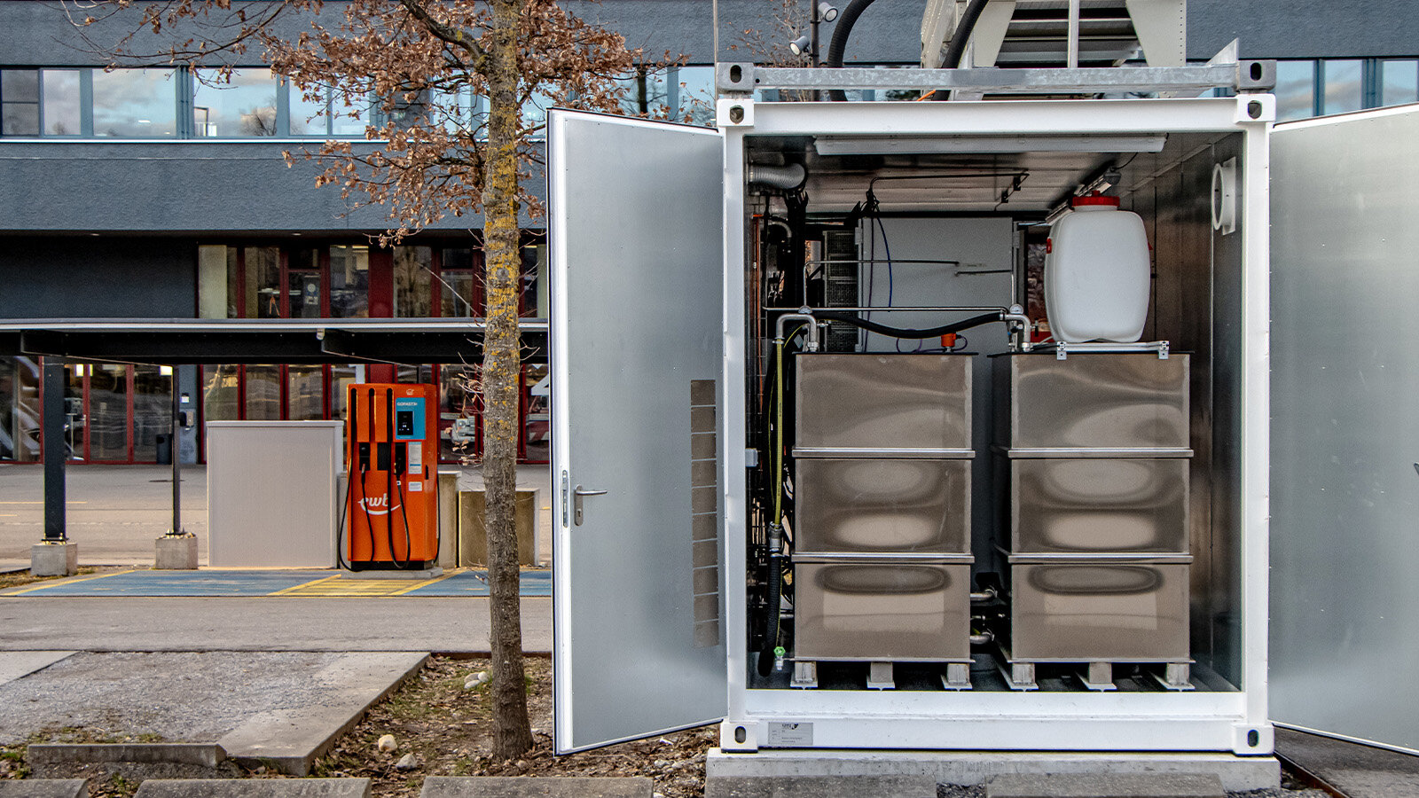 Auf dem Bild ist ein geöffneter Wasserstoffcontainer zu sehen, dessen Inhalt sichtbar ist. Im Hintergrund erkennt man einen Wohnblock oder ein Geschäftsgebäude.