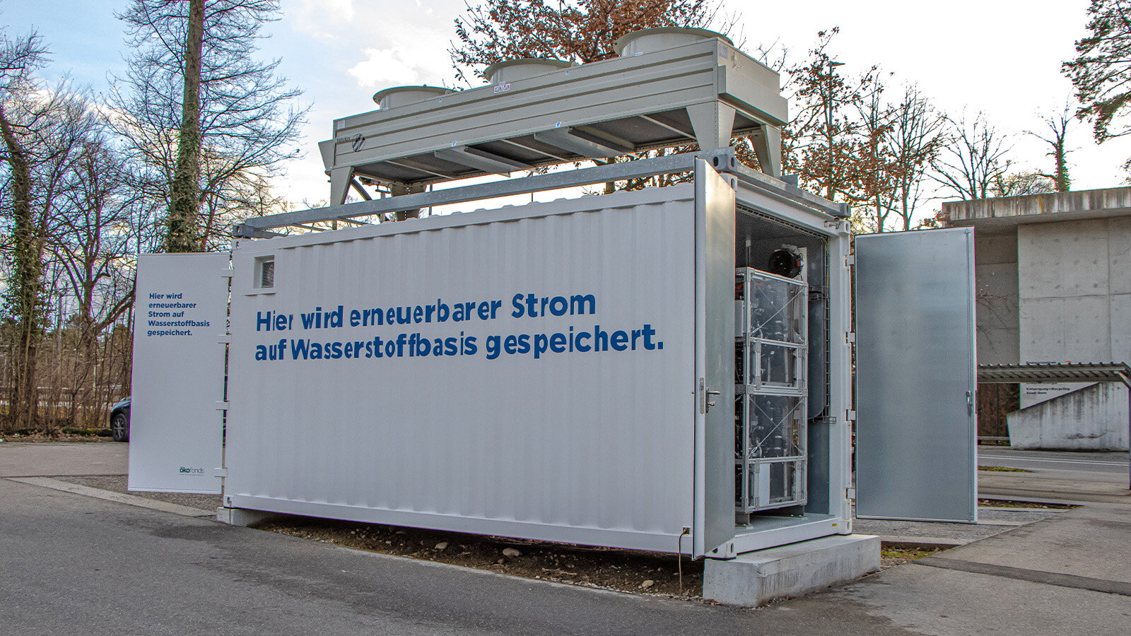 Auf dem Bild sieht man einen weißen Wasserstoffcontainer mit der blauen Aufschrift „Hier wird erneuerbarer Strom auf Wasserstoffbasis gespeichert“. Der Container ist quer dargestellt, und die rechte Tür steht offen. Im Hintergrund sind ein paar Bäume zu sehen.